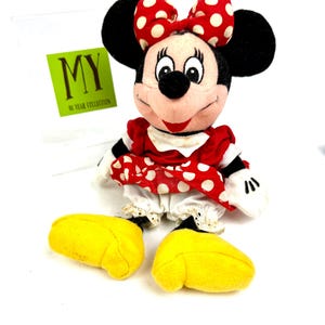 May include: A Minnie Mouse plush doll with a red and white polka dot bow, dress, and yellow shoes. The doll has black ears and a tag that reads "MY 90 YEAR COLLECTION".