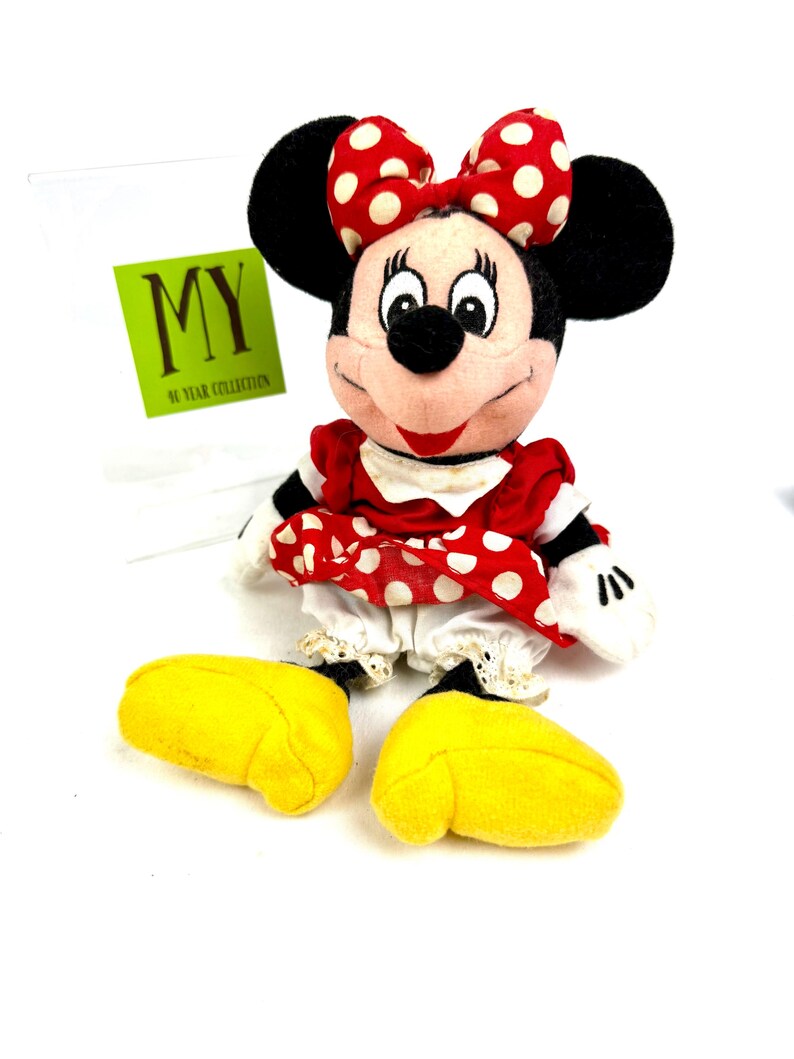 May include: A Minnie Mouse plush doll with a red and white polka dot bow, dress, and yellow shoes. The doll has black ears and a tag that reads "MY 90 YEAR COLLECTION".