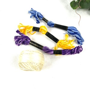 May include: Three spools of embroidery floss in purple, yellow, and blue, with a ball of white embroidery thread.