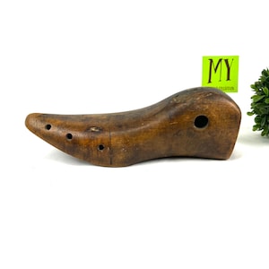 May include: A wooden shoe stretcher with a brown finish and multiple holes. The stretcher is shaped like a shoe and has a rounded toe.