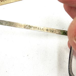 Pode incluir: Close-up de um par de &oacute;culos vintage. A arma&ccedil;&atilde;o de metal prateado apresenta o nome "ENDALL" e a medida "4 1/2" gravada na haste. O texto "PROTECT YOUR EYES... wear gogg" &eacute; vis&iacute;vel no fundo.