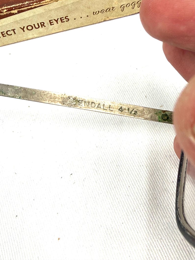 Pode incluir: Close-up de um par de &oacute;culos vintage. A arma&ccedil;&atilde;o de metal prateado apresenta o nome "ENDALL" e a medida "4 1/2" gravada na haste. O texto "PROTECT YOUR EYES... wear gogg" &eacute; vis&iacute;vel no fundo.
