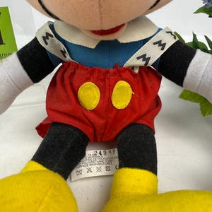 May include: A plush Mickey Mouse doll, featuring a blue shirt, red shorts with yellow buttons, and yellow shoes. The doll has black arms and legs, white gloves, and a smiling face. The doll has a tag with text.