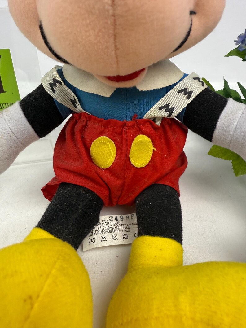 May include: A plush Mickey Mouse doll, featuring a blue shirt, red shorts with yellow buttons, and yellow shoes. The doll has black arms and legs, white gloves, and a smiling face. The doll has a tag with text.