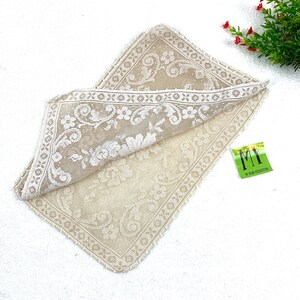 May include: A beige lace tablecloth with a floral pattern. The tablecloth is folded in half and has a small green tag with the text "MY 50 YEAR COLLECTION" on it.