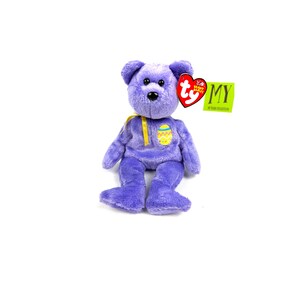May include: A lilac plush bear with a yellow ribbon and an Easter egg design on its chest. The bear has a red heart-shaped tag and a green tag that reads "MY".
