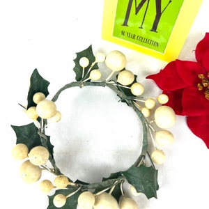 May include: A white and green Christmas candle ring with white berries and green holly leaves. The ring is made of wire and is decorated with faux berries and leaves.