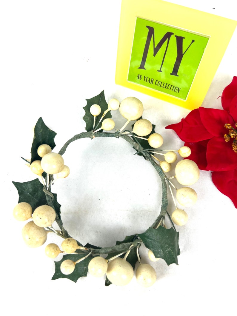 May include: A white and green Christmas candle ring with white berries and green holly leaves. The ring is made of wire and is decorated with faux berries and leaves.