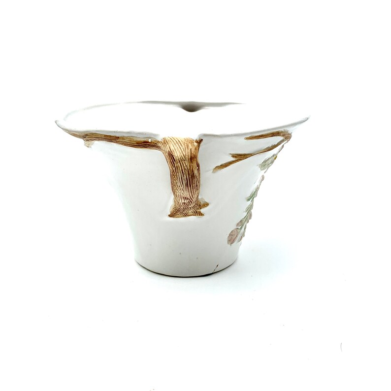 May include: White ceramic bowl with a flared rim and a textured, tree-like handle. The bowl features a brown and gold tree branch design on the side, with delicate floral accents in pink and green. The bowl has a unique, organic shape.