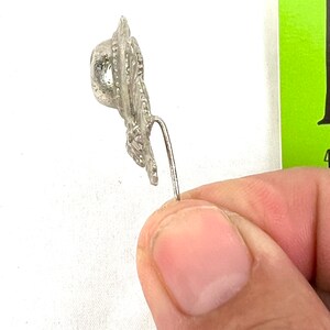 May include: A silver-toned metal pin with a detailed design resembling a hat. The pin has a long, thin stem.