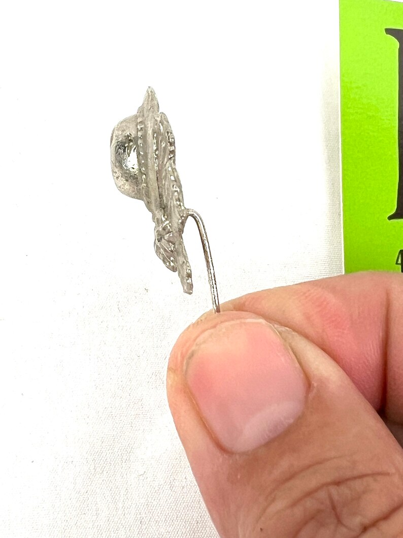 May include: A silver-toned metal pin with a detailed design resembling a hat. The pin has a long, thin stem.