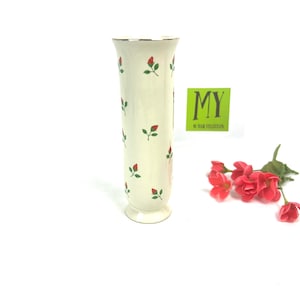 May include: A white ceramic vase with a gold rim and a pattern of red and green roses. The vase has a flared base and a cylindrical body.