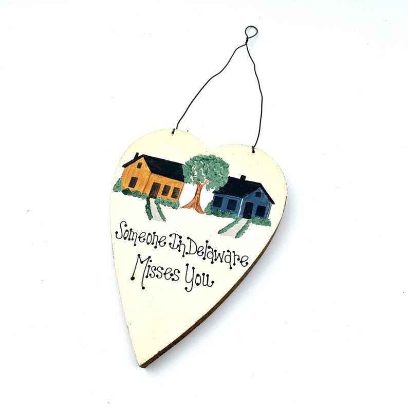 May include: A white heart-shaped wooden sign with a painted scene of two houses and a tree. The text "Someone in Delaware Misses You" is written in black cursive.