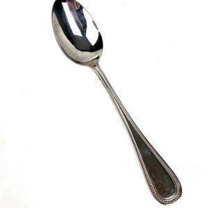 Op de afbeelding: Een zilveren lepel met een ovale kom en een lange steel. De steel heeft een decoratief randontwerp. De lepel is gemaakt van een glanzend, reflecterend metaal, waarschijnlijk roestvrij staal. De lepel staat tegen een witte achtergrond.