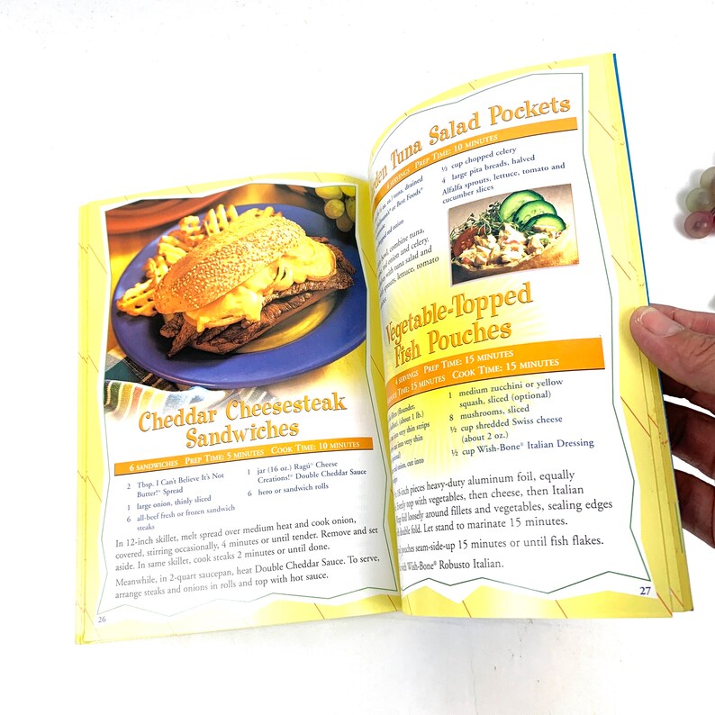 May include: A cookbook page with two recipes: Cheddar Cheesesteak Sandwiches and Vegetable-Topped Fish Pouches. The cheesesteak recipe includes ingredients like thinly sliced steak, cheese spread, and hero rolls. The fish recipe includes ingredients like zucchini, mushrooms, Swiss cheese, and Wish-Bone Italian Dressing.