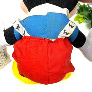 May include: A plush Mickey Mouse toy, viewed from the back. The toy features a black head, blue jacket, red shorts, and yellow shoes. White accents are visible on the collar and straps. The toy is designed with soft, plush materials.