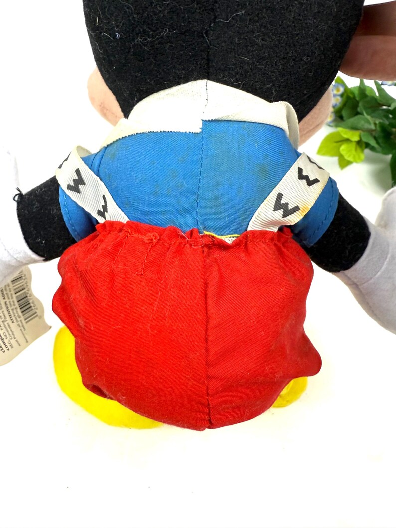 May include: A plush Mickey Mouse toy, viewed from the back. The toy features a black head, blue jacket, red shorts, and yellow shoes. White accents are visible on the collar and straps. The toy is designed with soft, plush materials.
