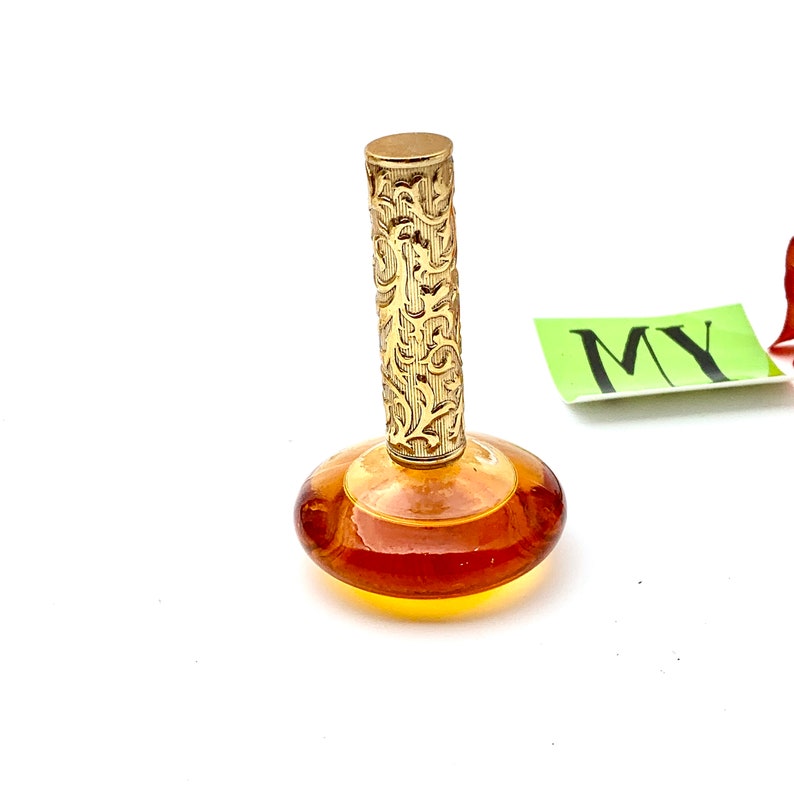May include: A vintage amber glass perfume bottle with a gold, ornate stopper. The bottle has a round, flat base and a cylindrical neck.