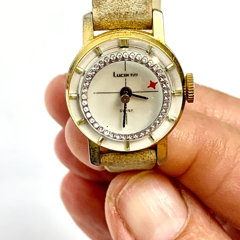 May include: A vintage gold-toned Lucetta Swiss watch with a cream-colored face. The watch face is encircled with small, clear stones. The watch has a gold-toned bezel and a worn, light-tan leather band.