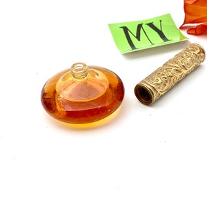 May include: A vintage amber glass perfume bottle with a gold-colored metal applicator. The bottle has a round, flat base and a clear glass top. The applicator has an intricate design.