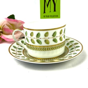 May include: A white teacup and saucer with a green and gold leaf design. The saucer has a gold rim and the teacup has a gold band around the top. The teacup has a small handle.