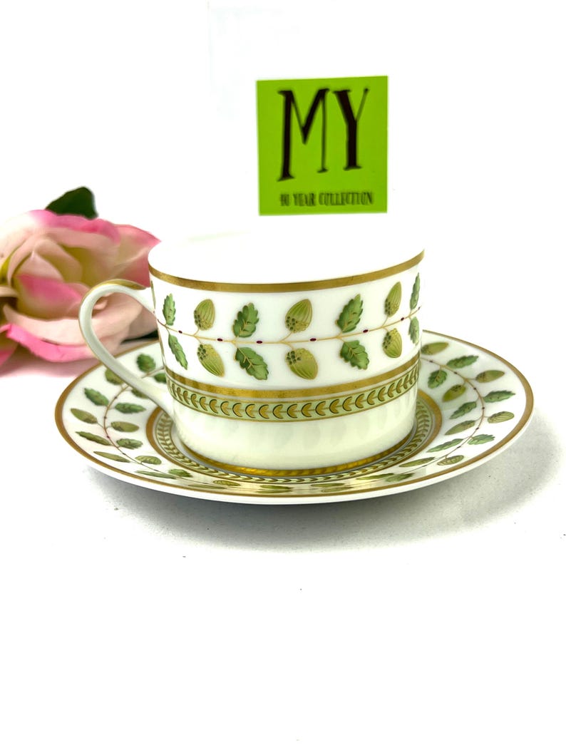 May include: A white teacup and saucer with a green and gold leaf design. The saucer has a gold rim and the teacup has a gold band around the top. The teacup has a small handle.
