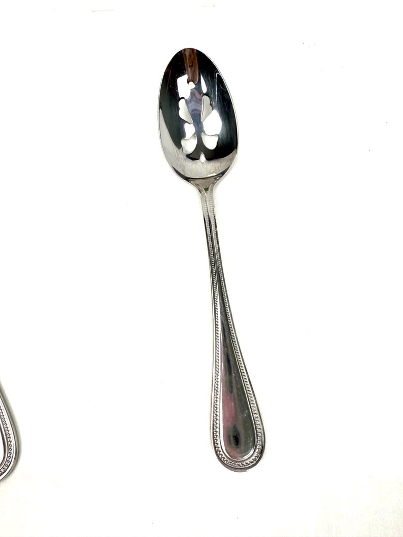 Op de afbeelding: Een zilveren serveerlepel met een sleufkom met een decoratief bloemmotief. De steel heeft een kralenrand en een druppelvormig uiteinde. De lepel is ontworpen om te serveren en is gemaakt van een glanzend, reflecterend metaal.