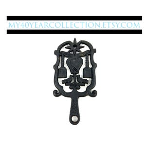 May include: Black cast iron hot pad with a heart and two stylised brooms in the centre. The hot pad has a decorative handle.