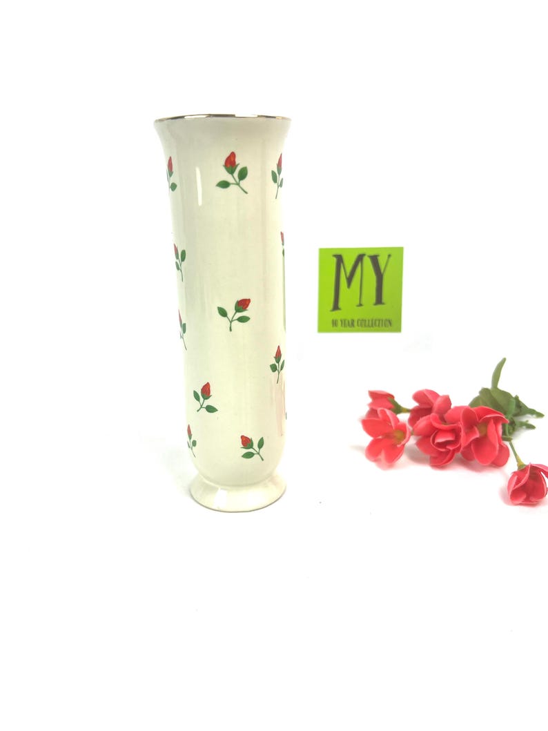 May include: A white ceramic vase with a gold rim and a pattern of red roses and green leaves. The vase is about 10 inches tall and has a round base.
