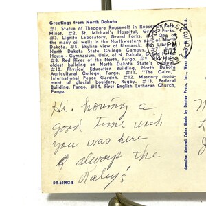 May include: A vintage postcard with a blue border featuring a list of attractions in North Dakota. The postcard has a handwritten message that reads "Hi. houng a good time wish you was here & alwoup the Nalys".