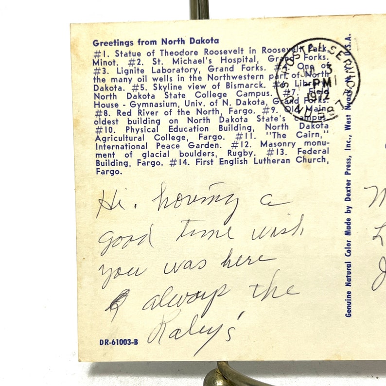 May include: A vintage postcard with a blue border featuring a list of attractions in North Dakota. The postcard has a handwritten message that reads "Hi. houng a good time wish you was here & alwoup the Nalys".