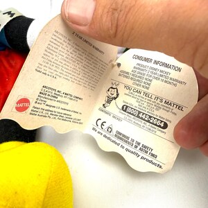 May include: A Mickey Mouse plush toy with a yellow body, red shorts, and black ears. The toy has a tag with consumer information, including a 2-year limited warranty and safety compliance details.