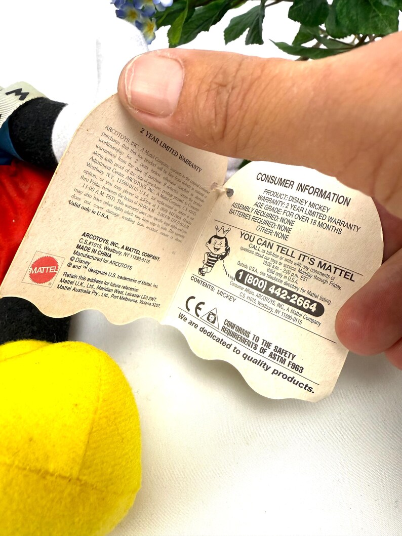 May include: A Mickey Mouse plush toy with a yellow body, red shorts, and black ears. The toy has a tag with consumer information, including a 2-year limited warranty and safety compliance details.