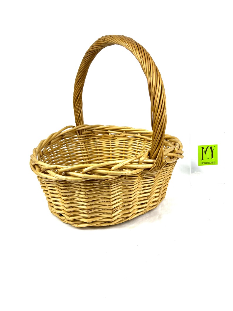 May include: A natural woven wicker basket with a rounded oval shape and a single handle. The basket is light brown in color and has a woven pattern.