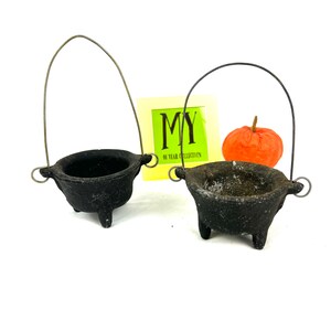 May include: Two small black cast iron cauldrons with wire handles. The cauldrons are on a white background.