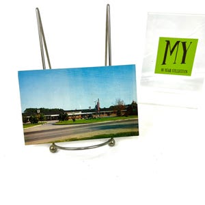 May include: A silver metal postcard holder with a postcard featuring a photo of a building with a blue sky and green grass.