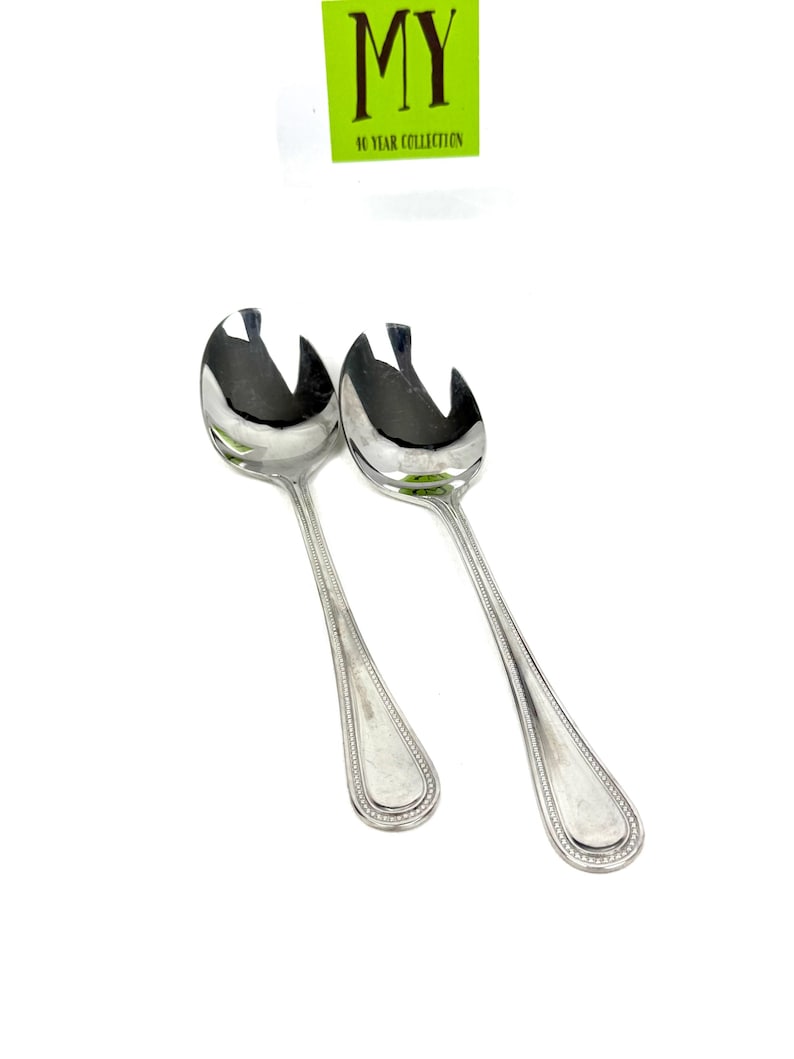 May include: Two shiny, silver serving spoons with a unique slotted design. The spoons have long, slender handles with a beaded border and a teardrop-shaped end. The image includes a green and white tag that reads "MY 10 YEAR COLLECTION."
