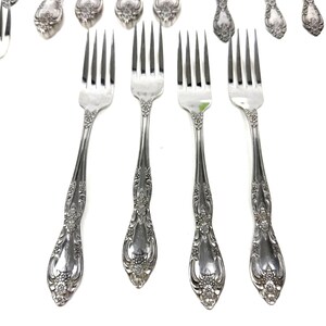 May include: Silverware set featuring ornate forks and spoons with floral detailing. The forks are in the foreground, with tines pointing upwards. The spoons and additional forks are in the background, all on a white background.
