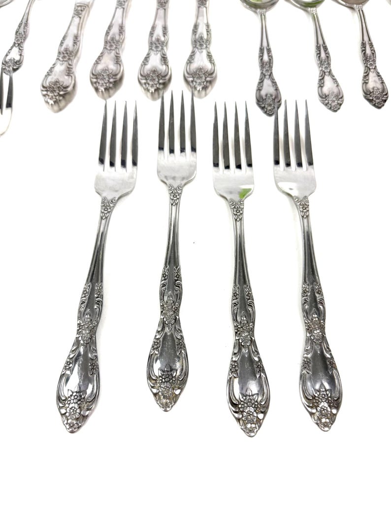 May include: Silverware set featuring ornate forks and spoons with floral detailing. The forks are in the foreground, with tines pointing upwards. The spoons and additional forks are in the background, all on a white background.