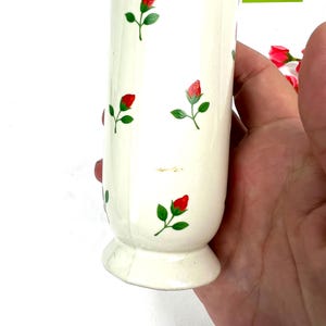 May include: White ceramic vase with a floral pattern of red roses and green leaves. The vase has a flared base and a cylindrical body. The vase is decorated with a small tag that reads "MY 10 YEAR COLLECTION".
