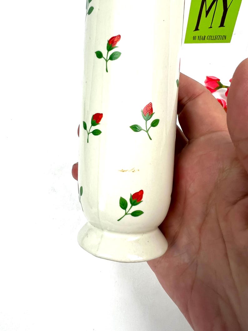 May include: White ceramic vase with a floral pattern of red roses and green leaves. The vase has a flared base and a cylindrical body. The vase is decorated with a small tag that reads "MY 10 YEAR COLLECTION".