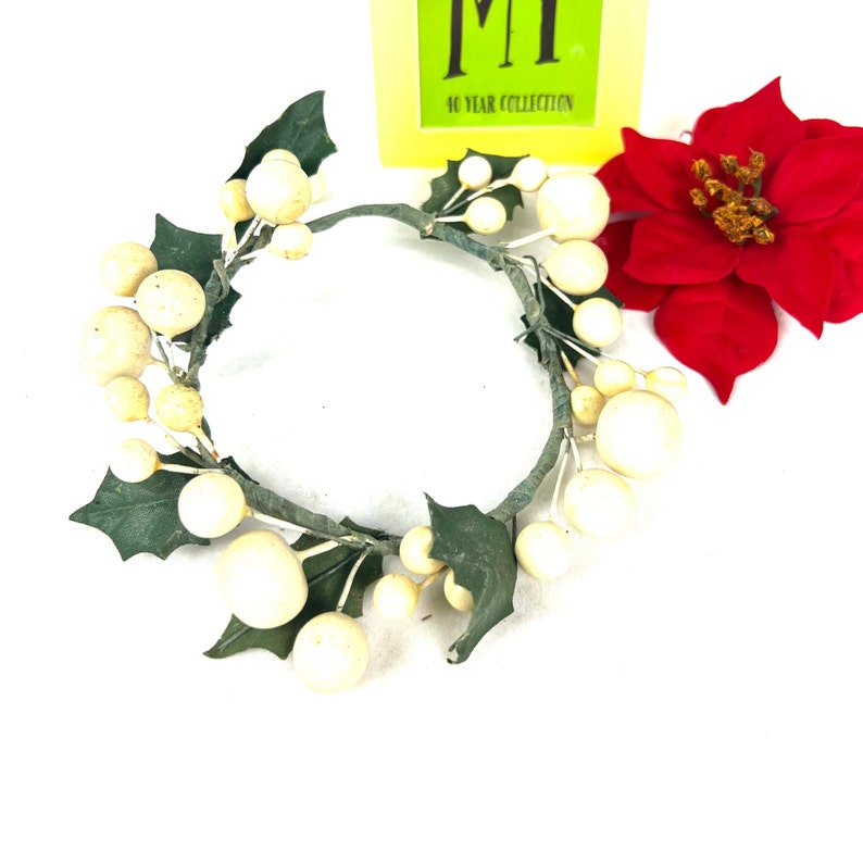 May include: A white, artificial holly wreath with green leaves and white berries. The wreath is made of wire and is suitable for decorating a candle or other small object.
