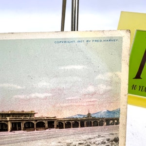 May include: A vintage postcard with a colour image of a building with arched openings, set against a backdrop of mountains and a cloudy sky. The text "COPYRIGHT, 1907, BY FRED. HARVEY" is printed at the top. A green and yellow card is in the background.