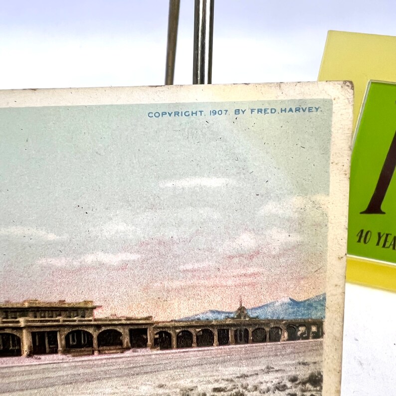 May include: A vintage postcard with a colour image of a building with arched openings, set against a backdrop of mountains and a cloudy sky. The text "COPYRIGHT, 1907, BY FRED. HARVEY" is printed at the top. A green and yellow card is in the background.