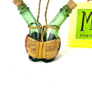 May include: Two small green glass bottles with cork tops are tied together with twine and placed in a woven basket. The basket is hanging from a twine loop.