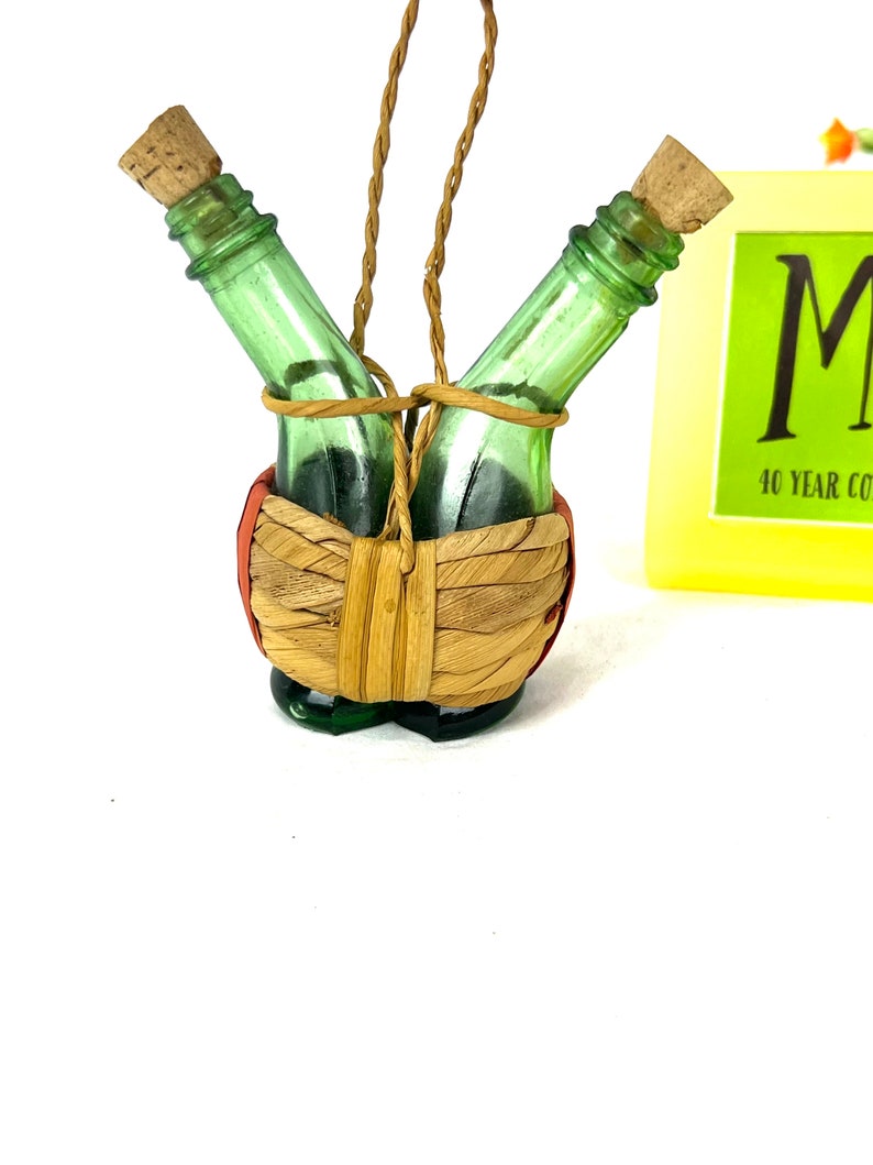 May include: Two small green glass bottles with cork tops are tied together with twine and placed in a woven basket. The basket is hanging from a twine loop.