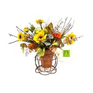 Könnte beinhalten: Ein herbstliches Blumenarrangement mit Sonnenblumen, künstlichen Kürbissen und grünen Blättern in einem braunen Terrakotta-Topf mit einem Drahtgestell. Das Arrangement ist mit "MY HOME COLLECTION" beschriftet.