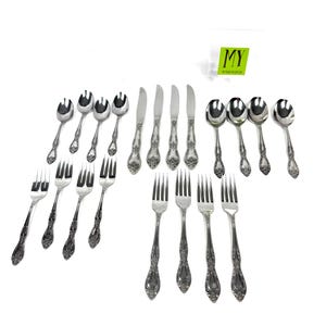 May include: A collection of ornate silver-colored flatware, including forks, knives, and spoons, arranged on a white surface. The silverware features detailed floral patterns on the handles. The image includes a small green and white tag with the letters "MY".