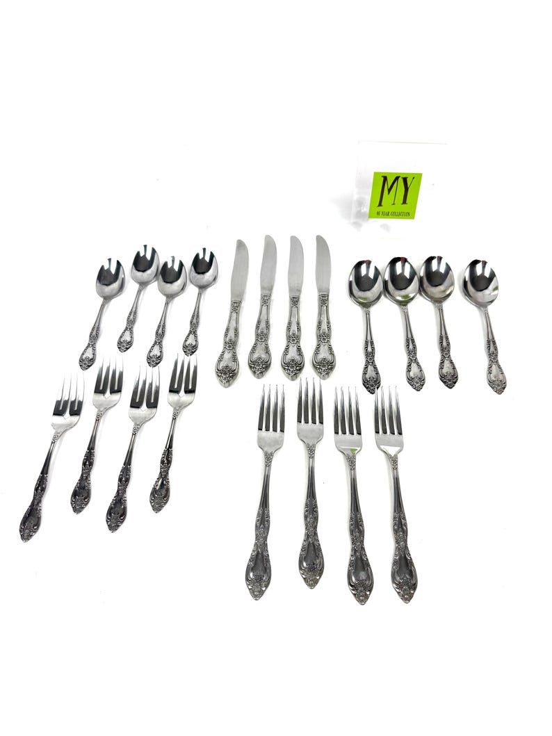 May include: A collection of ornate silver-colored flatware, including forks, knives, and spoons, arranged on a white surface. The silverware features detailed floral patterns on the handles. The image includes a small green and white tag with the letters "MY".