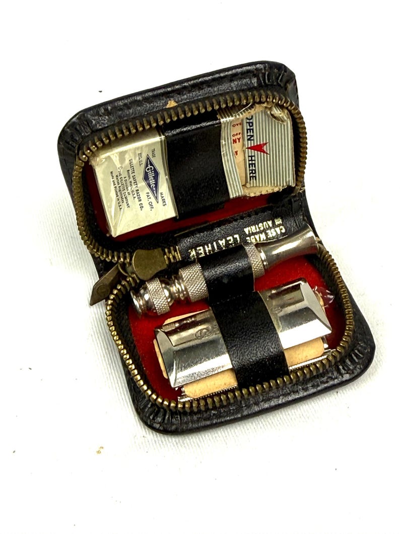 May include: Vintage black leather travel shaving kit with a zipper closure. The kit contains a Gillette safety razor, blades, and a small strop. The interior is lined with red fabric. The case is marked "CASE MADE LEATHER AUSTRIA".